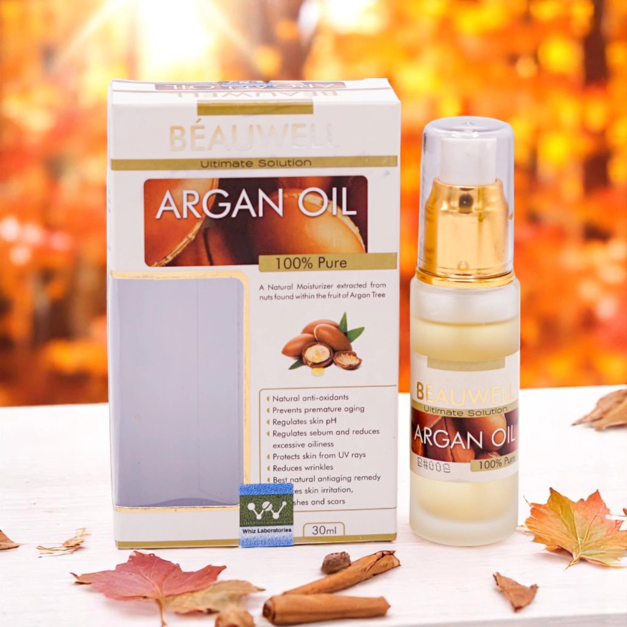 Beauwell Ultimate Solution Argan Oil 100% Pure, 30ml clear pump bottle with a gold top and a white and gold label, and its matching white and gold box, set against a background of autumn leaves and cinnamon sticks.