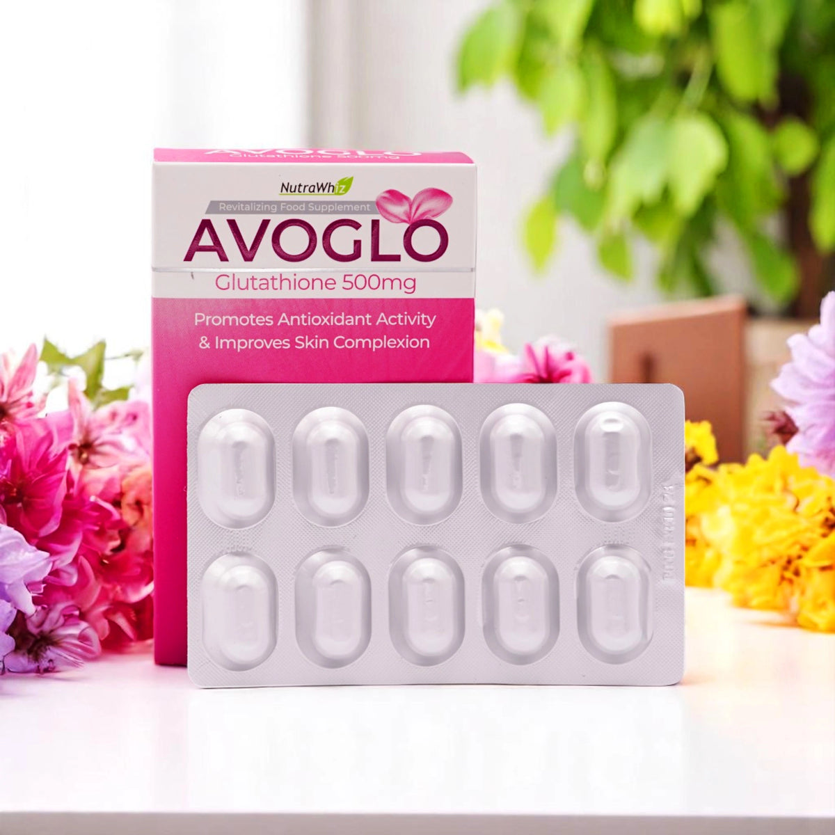 Nutrawhiz Avoglo Glutathione 500mg capsules, a blister pack of 10 capsules with a white and pink box, set among pink, yellow, and purple flowers on a white surface.