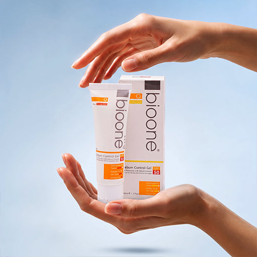 Two hands holding a tube and box of bioone Vitamin Control Gel SPF 50 against a soft light blue gradient background.