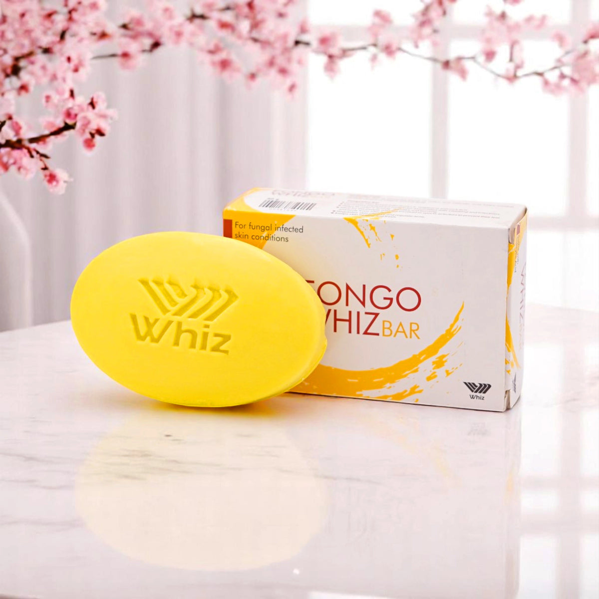 Fongo Whiz Bar, an oval-shaped yellow soap with 'Whiz' embossed on it, and its white and orange box, set on a white marble surface with pink cherry blossoms in the background.