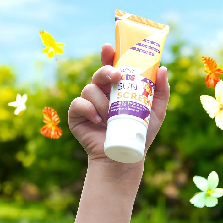A close-up of a child's hand holding a tube of Whiz Kids Sunscreen in a bright outdoor setting with floating butterflies.