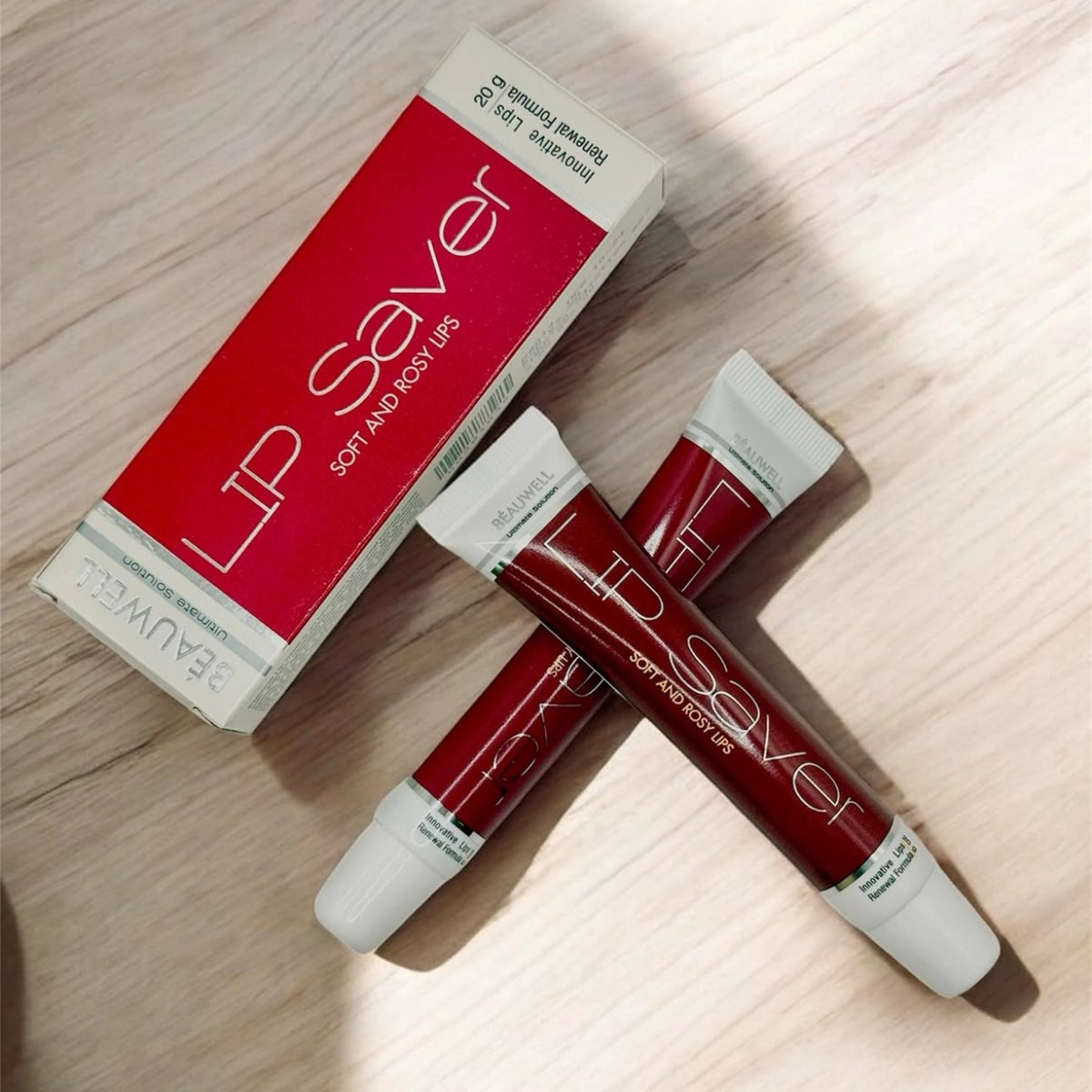 A flat lay photo of two Beauwell Lip Saver cream tubes crossed over each other on a wooden surface, next to their red and white box.
