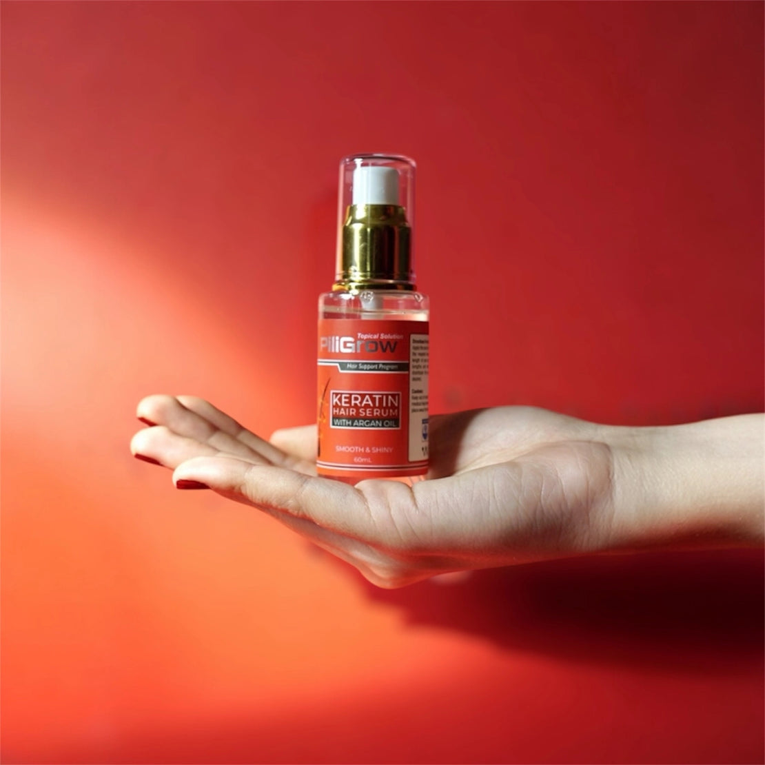 A hand holds a clear bottle of PiliGrow Keratin Hair Serum with a gold pump dispenser against a red background.