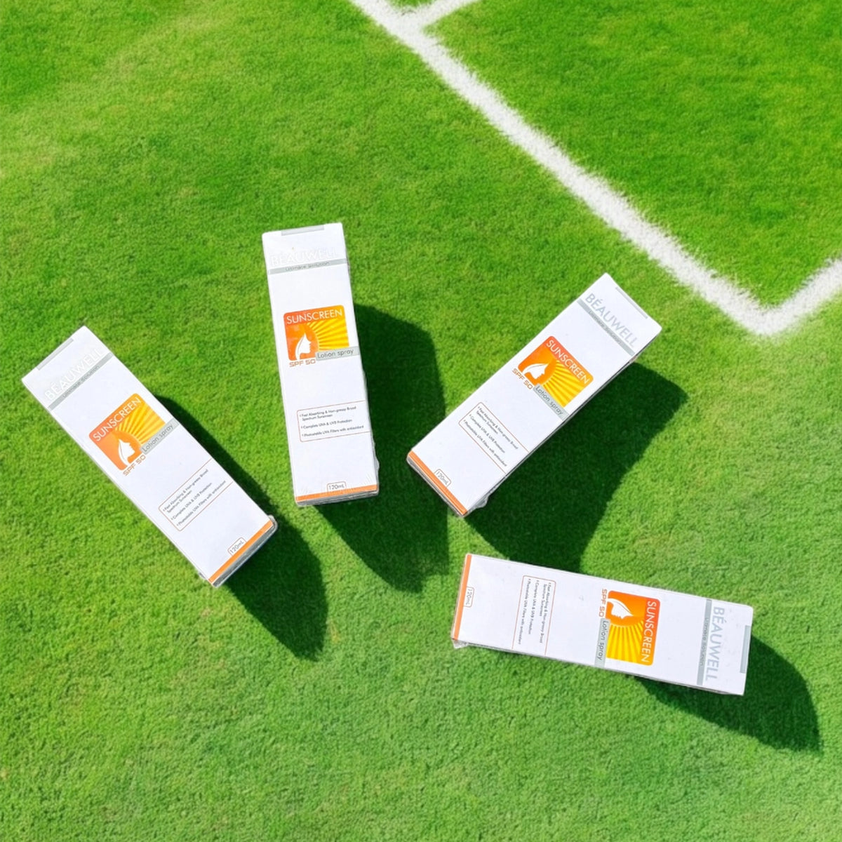 An overhead shot of four white and orange boxes of Beauwell Sunscreen Lotion Spray lying on a green surface that resembles a ground.